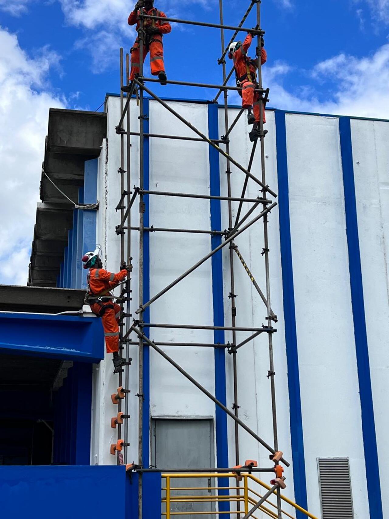 Quatro IRATAs subindo andaime em fachada industrial azul